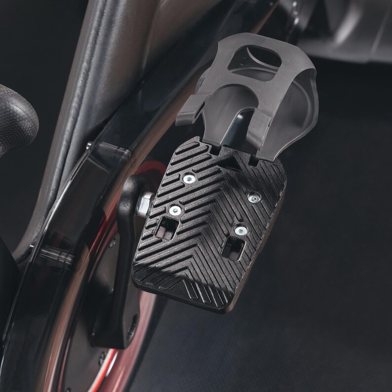 May include: Close-up of a black exercise bike pedal with a textured surface and a foot cage. The pedal is attached to the bike's crank arm and is designed for secure foot placement during workouts. The bike frame is visible in the background.