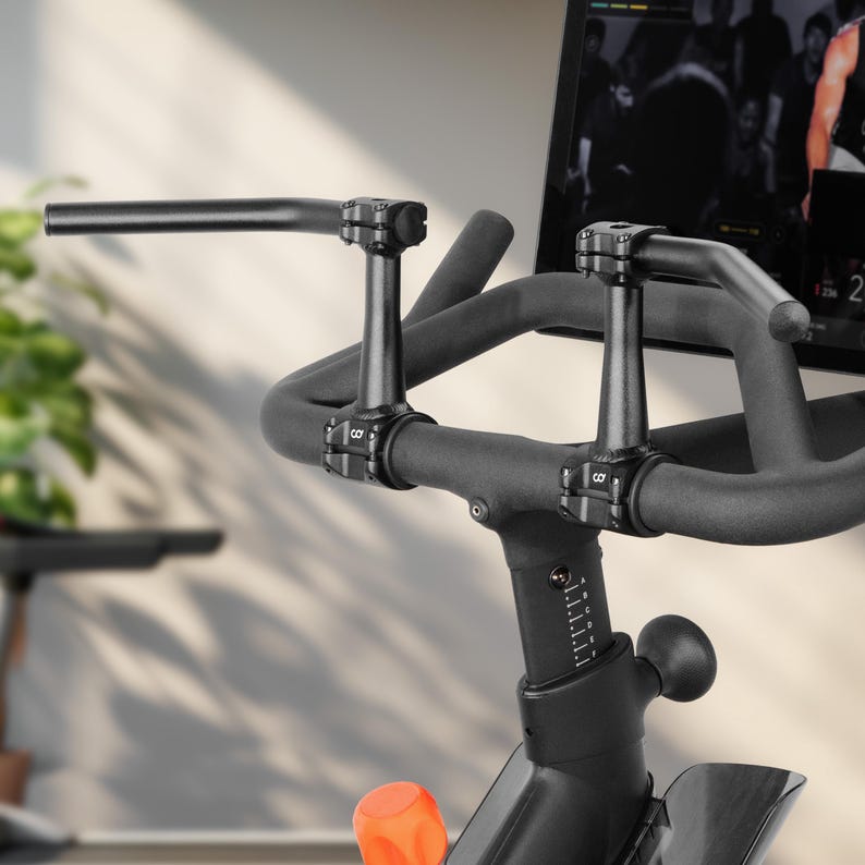Puede incluir: Primer plano de una bicicleta est&aacute;tica negra. El manillar es negro con un dise&ntilde;o curvo y empu&ntilde;aduras acolchadas. La bicicleta tiene una perilla de ajuste naranja y una pantalla digital en el fondo.