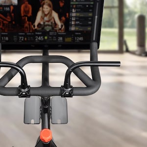 May include: Indoor exercise bike with a large screen displaying a cycling class. The bike has a black frame, handlebars, and a screen. The screen shows a woman leading a class. The bike is in a bright, modern gym setting.