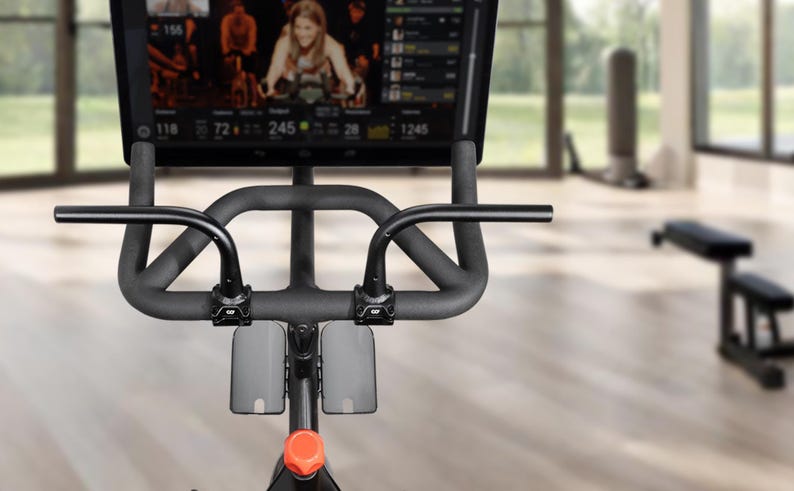 May include: Indoor exercise bike with a large screen displaying a cycling class. The bike has a black frame, handlebars, and a screen. The screen shows a woman leading a class. The bike is in a bright, modern gym setting.