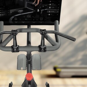 Puede incluir: Primer plano de una bicicleta est&aacute;tica con pantalla digital. La bicicleta tiene un manillar gris y un bot&oacute;n de ajuste rojo. La pantalla muestra m&eacute;tricas de entrenamiento. Una cinta de correr est&aacute; borrosa en el fondo.