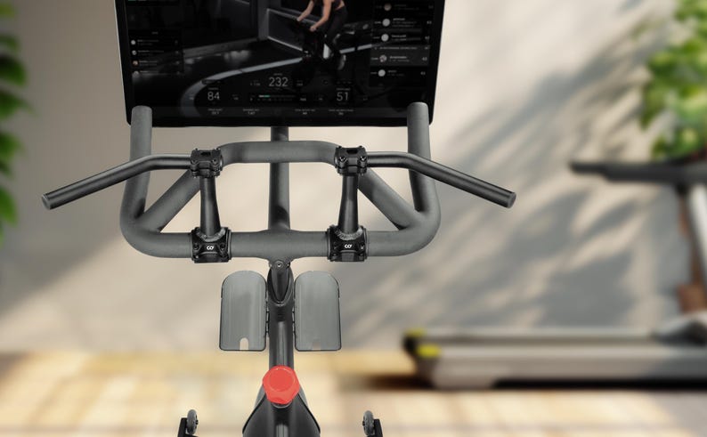 Puede incluir: Primer plano de una bicicleta est&aacute;tica con pantalla digital. La bicicleta tiene un manillar gris y un bot&oacute;n de ajuste rojo. La pantalla muestra m&eacute;tricas de entrenamiento. Una cinta de correr est&aacute; borrosa en el fondo.