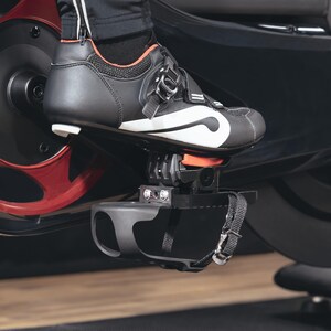 May include: Close-up of a black and white cycling shoe secured to a black pedal. The shoe has a white stripe and orange accents. The pedal is attached to a stationary bike with a red and black flywheel.