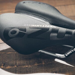 May include: Close-up of two black bicycle seats. The top seat is slightly out of focus, while the front seat displays the brand name "OZO VENTURE evo" in a dark gray color. A silver bar with measurement markings is visible.
