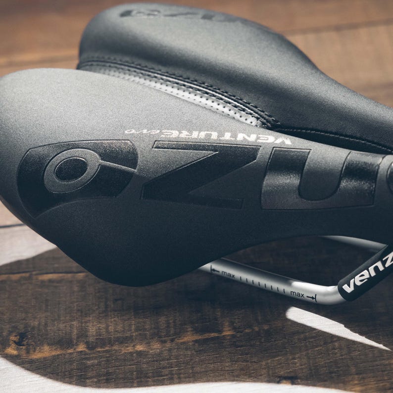 May include: Close-up of two black bicycle seats. The top seat is slightly out of focus, while the front seat displays the brand name "OZO VENTURE evo" in a dark gray color. A silver bar with measurement markings is visible.