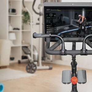 May include: Indoor exercise bike with a large screen displaying a workout session. The bike has black handlebars and a red adjustment knob. A gray exercise ball and blue yoga mat are in the foreground, suggesting a home gym setup.