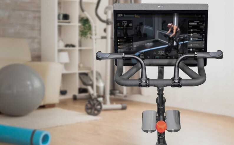 May include: Indoor exercise bike with a large screen displaying a workout session. The bike has black handlebars and a red adjustment knob. A gray exercise ball and blue yoga mat are in the foreground, suggesting a home gym setup.