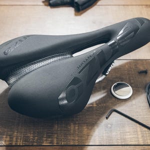 May include: A black bicycle saddle with the word "VENTURE" printed on the side. The saddle is surrounded by bicycle parts, including a small Allen wrench, a round white and silver object, and a black plastic piece. The items are on a wooden surface.