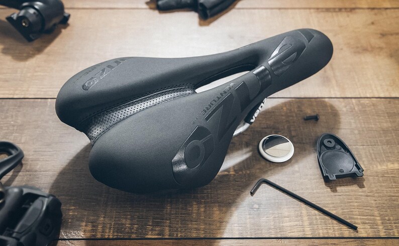 May include: A black bicycle saddle with the word "VENTURE" printed on the side. The saddle is surrounded by bicycle parts, including a small Allen wrench, a round white and silver object, and a black plastic piece. The items are on a wooden surface.