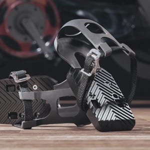 May include: Close-up of two black and gray exercise bike pedals with adjustable straps and a textured surface. The pedals are on a wooden surface, with a blurred exercise bike in the background. Small screws and tools are also visible.