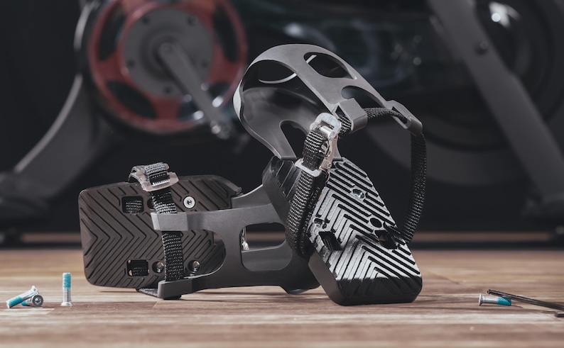 May include: Close-up of two black and gray exercise bike pedals with adjustable straps and a textured surface. The pedals are on a wooden surface, with a blurred exercise bike in the background. Small screws and tools are also visible.