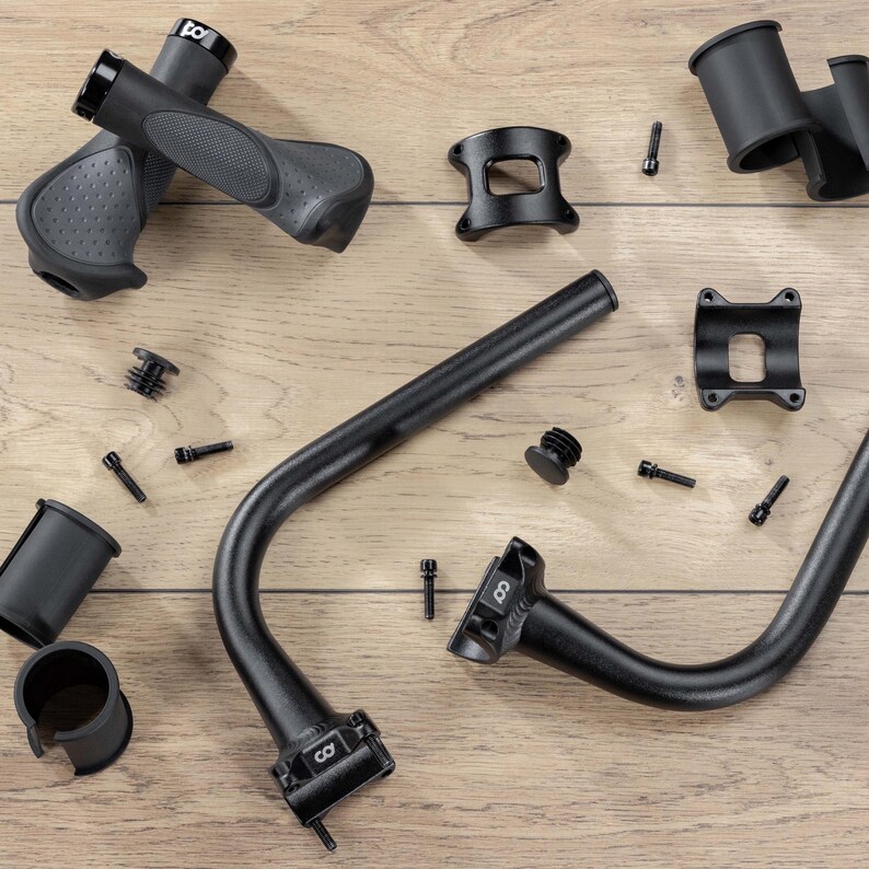 May include: An assortment of black bicycle parts, including handlebars, grips, stem, and various small screws and plugs. The parts are arranged on a light brown wooden surface, showcasing the components' design and texture.