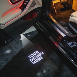Puede incluir: Interior de puerta de coche con panel negro, asiento de cuero beige y alfombra negra. Una luz blanca proyecta el texto "YOUR DESIGN HERE" sobre la alfombra. Iluminación ambiental roja visible.