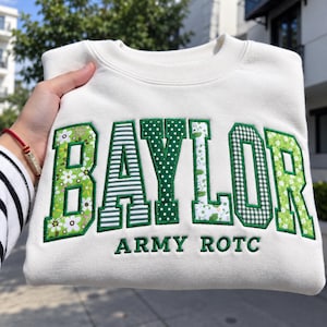 Könnte beinhalten: Ein cremefarbenes Sweatshirt mit Rundhalsausschnitt und dem Wort BAYLOR in großen Blockbuchstaben. Jeder Buchstabe ist mit einem anderen grün-weißen Muster gefüllt, darunter Blumen-, Streifen- und Punktedesigns. Darunter ist ARMY ROTC gestickt.