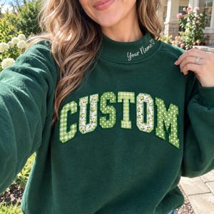 May include: A dark green mock neck sweatshirt featuring the word CUSTOM in large, block-style letters across the chest. The letters are filled with a mix of green gingham, white polka dot, and yellow floral patterns. The text Your Name is embroidered.