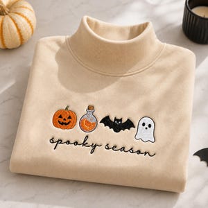 May include: A beige mock neck sweatshirt featuring embroidered Halloween icons including an orange jack-o'-lantern, a potion bottle, a black bat, and a white ghost, with the black cursive text "spooky season" embroidered below the designs.