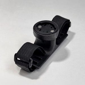 May include: Black bicycle computer mount with adjustable straps. The mount has a circular base with two screws and a textured surface. The straps are made of a woven material and have a buckle closure. The mount is designed to hold a bicycle computer.