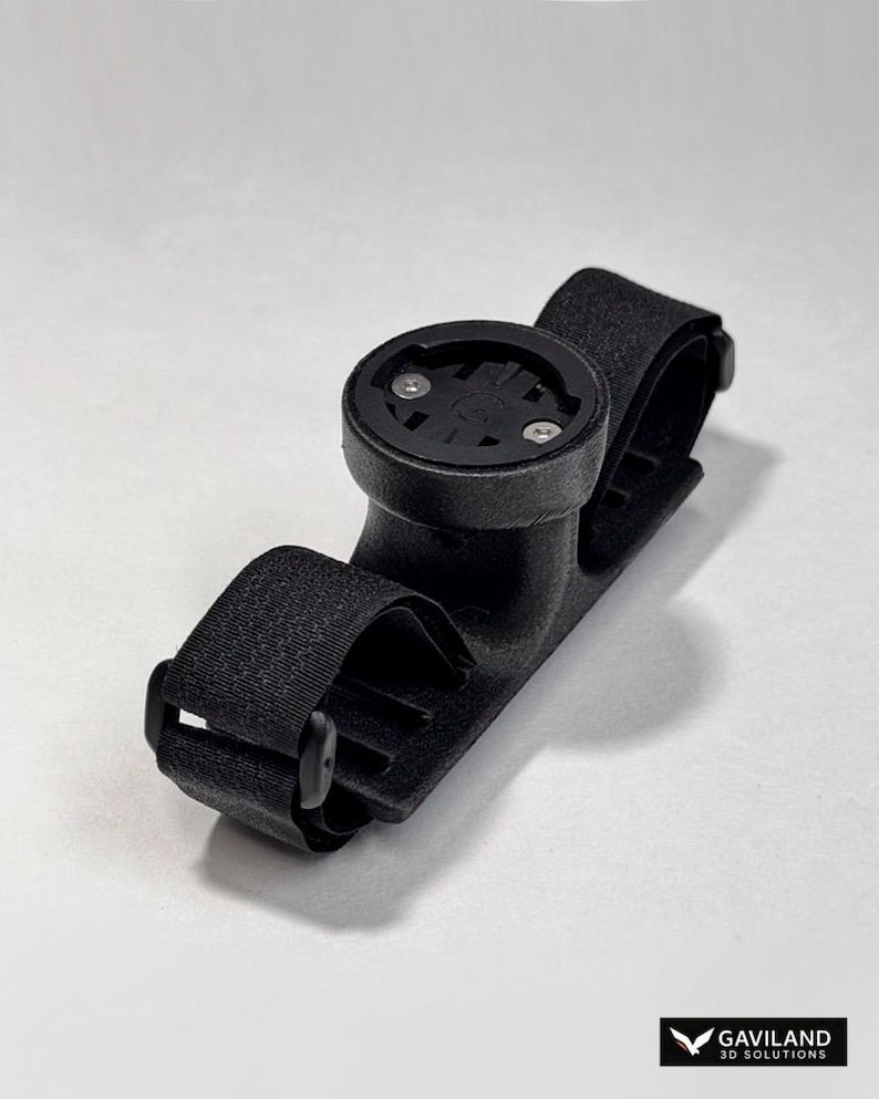 May include: Black bicycle computer mount with adjustable straps. The mount has a circular base with two screws and a textured surface. The straps are made of a woven material and have a buckle closure. The mount is designed to hold a bicycle computer.