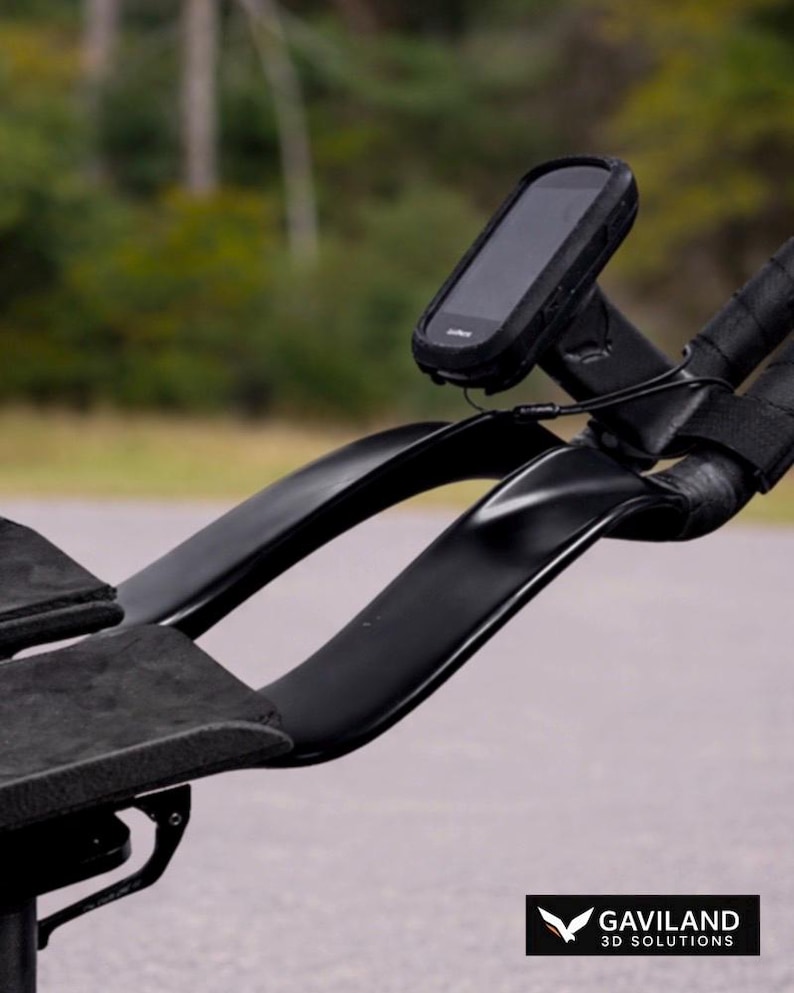 May include: Black bicycle handlebars with a mounted black device, possibly a GPS or cycling computer. The handlebars have integrated armrests with black padding. The image is set against a blurred outdoor background.