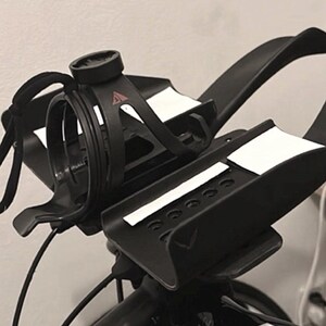 May include: Black bicycle handlebar with a water bottle cage and a computer mount. The handlebar has white accents and is attached to a black bicycle frame. The image is a close-up shot.