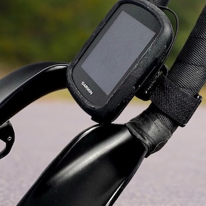 May include: Black Garmin cycling computer mounted on a bicycle handlebar. The computer is in a black protective case and secured with a black strap. The handlebar is black and has a textured grip. The Garmin logo is visible.