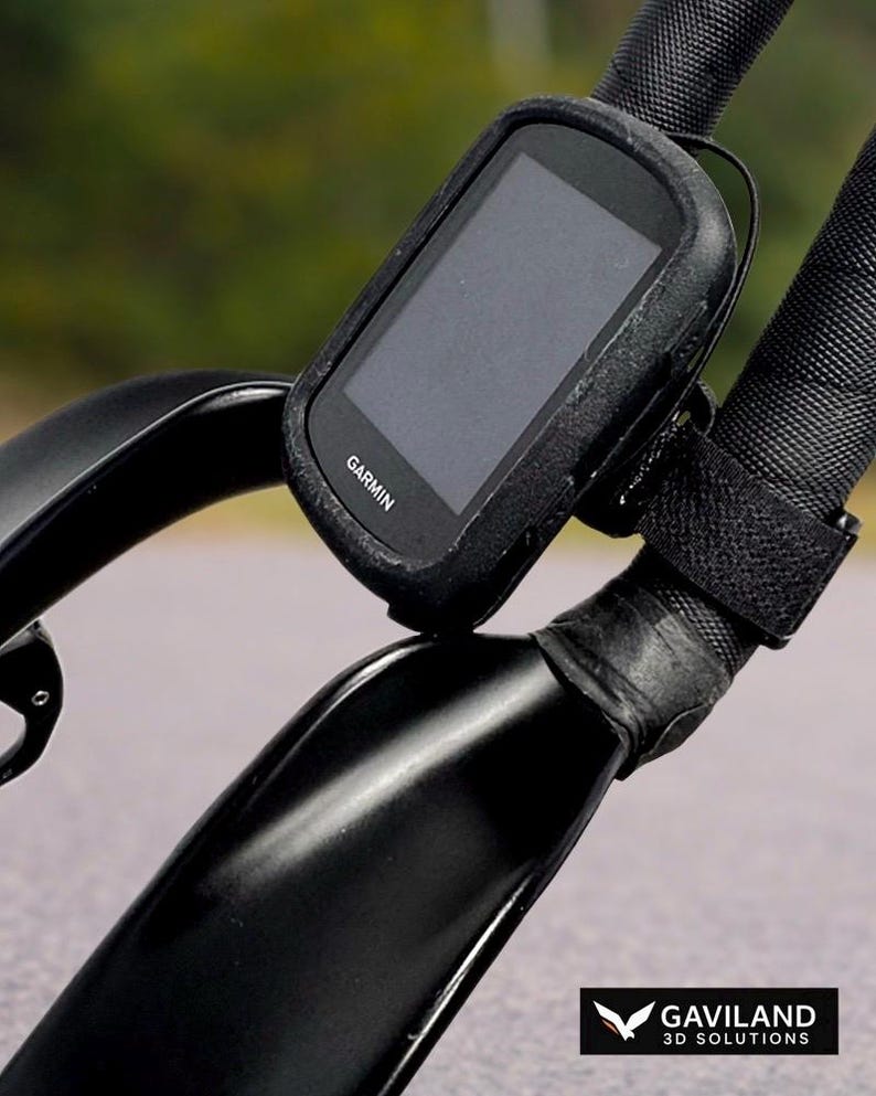 May include: Black Garmin cycling computer mounted on a bicycle handlebar. The computer is in a black protective case and secured with a black strap. The handlebar is black and has a textured grip. The Garmin logo is visible.