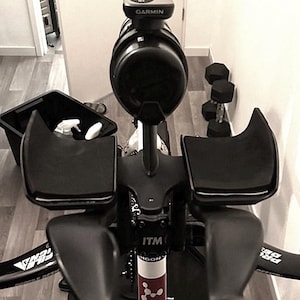 May include: Black and white indoor exercise bike with a Garmin device and black armrests. The bike has a black frame and is set up on a wooden floor. Dumbbells are visible in the background.