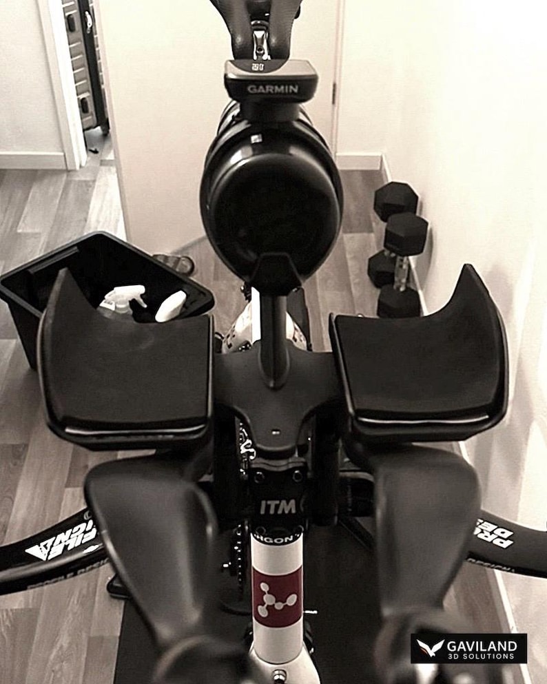 May include: Black and white indoor exercise bike with a Garmin device and black armrests. The bike has a black frame and is set up on a wooden floor. Dumbbells are visible in the background.