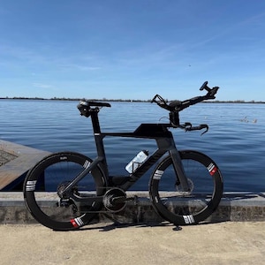 May include: A black triathlon bicycle with aerodynamic wheels and a water bottle, positioned near a body of water under a blue sky. The bike has a unique handlebar design and is designed for speed and efficiency.