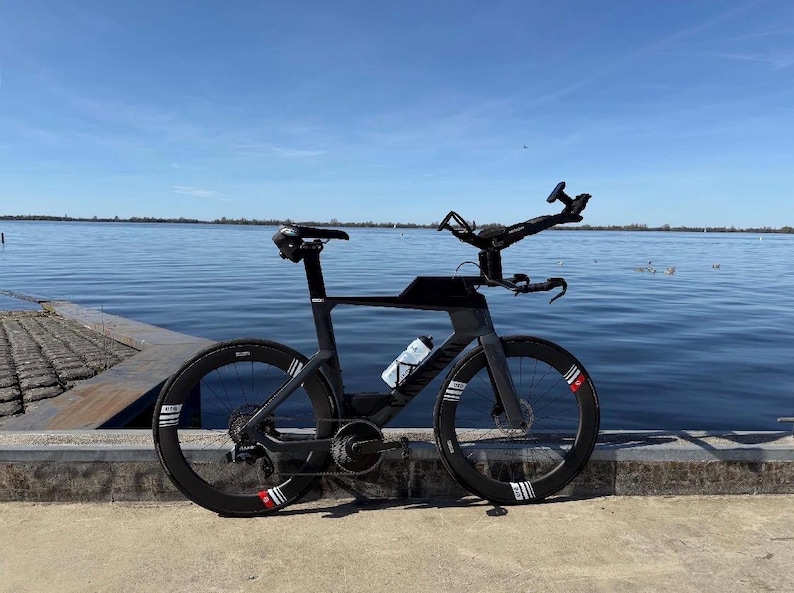 May include: A black triathlon bicycle with aerodynamic wheels and a water bottle, positioned near a body of water under a blue sky. The bike has a unique handlebar design and is designed for speed and efficiency.