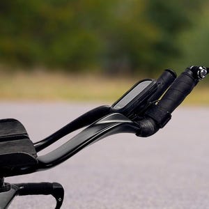 May include: Black triathlon bike handlebars with a mounted computer and wrapped grips. The handlebars curve upwards and have a sleek, aerodynamic design. The bike is on a paved road with a blurred green background.