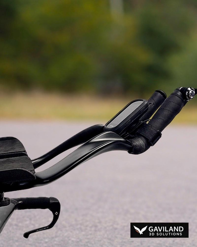 May include: Black triathlon bike handlebars with a mounted computer and wrapped grips. The handlebars curve upwards and have a sleek, aerodynamic design. The bike is on a paved road with a blurred green background.