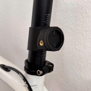 May include: A black 3D-printed mount attached to a bicycle seat post. The mount features a circular interface with a brass threaded insert on the side. The seat post is black with white measurement markings, secured by a metal clamp to a white frame.