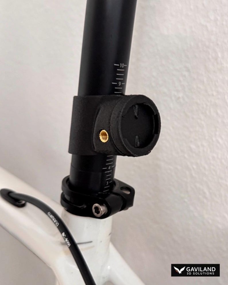 May include: A black 3D-printed mount attached to a bicycle seat post. The mount features a circular interface with a brass threaded insert on the side. The seat post is black with white measurement markings, secured by a metal clamp to a white frame.