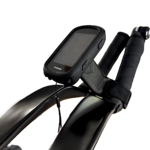 May include: Black Garmin cycling computer mounted on black bicycle handlebars. The computer is in a black protective case. The handlebars are wrapped in black tape. The image is set against a white background.