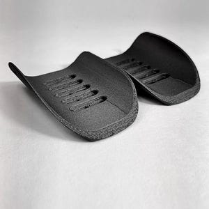 May include: Two black, curved, semi-cylindrical guards with a textured surface and a series of parallel vertical slots along the center. The objects feature a matte finish and are positioned side-by-side. A logo reads GAVILAND 3D SOLUTIONS.