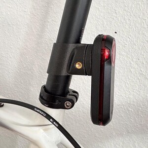 May include: A black bicycle taillight mounted to a black seat post on a white bicycle frame. The light features a vertical red lens and a brass-colored screw insert on the side of the mounting bracket. A black Shimano BC-9000 cable is visible.