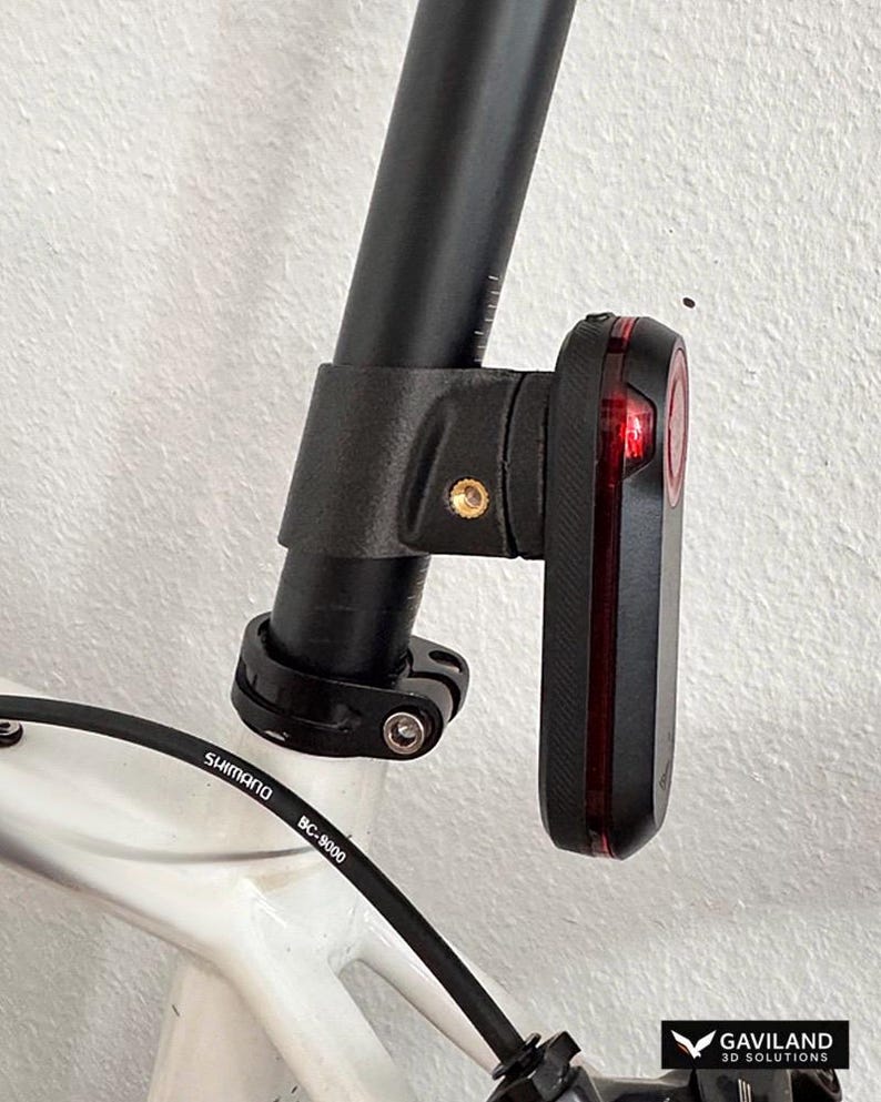 May include: A black bicycle taillight mounted to a black seat post on a white bicycle frame. The light features a vertical red lens and a brass-colored screw insert on the side of the mounting bracket. A black Shimano BC-9000 cable is visible.