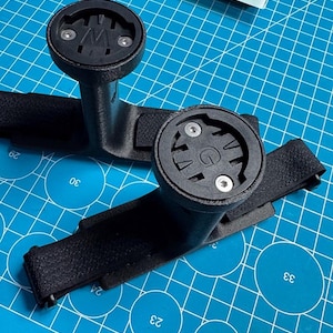 May include: Two black bicycle computer mounts with a strap and a circular top. The mounts are on a blue cutting mat. The logo "GAVILAND 3D SOLUTIONS" is in the bottom right corner.