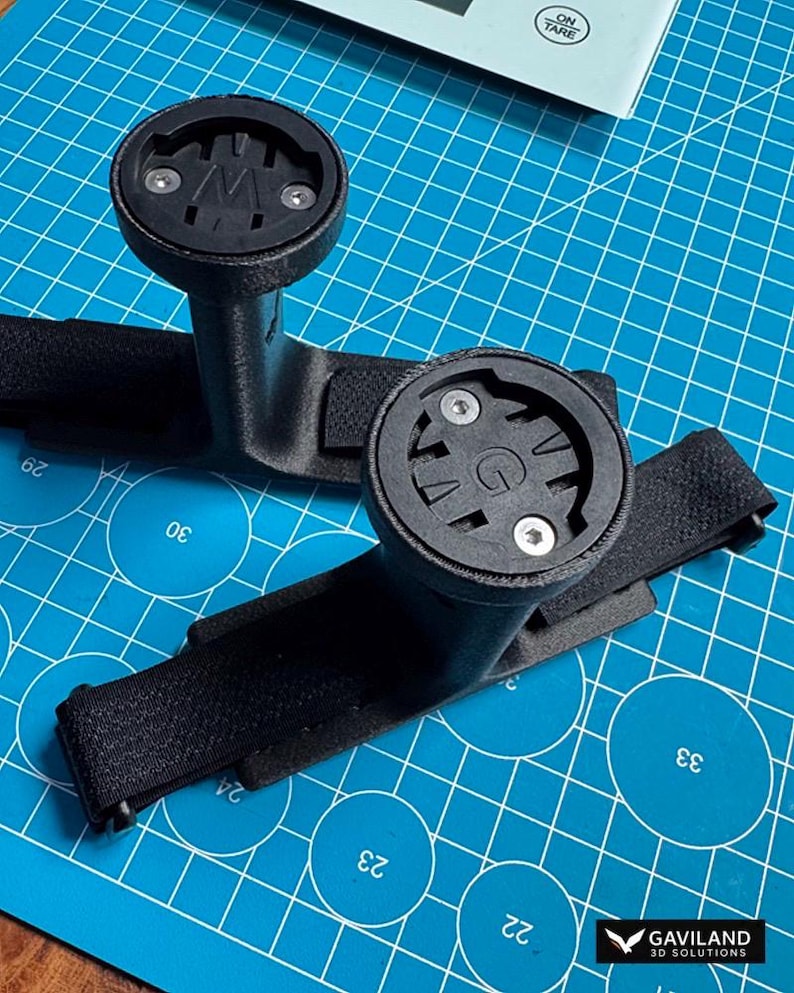 May include: Two black bicycle computer mounts with a strap and a circular top. The mounts are on a blue cutting mat. The logo "GAVILAND 3D SOLUTIONS" is in the bottom right corner.