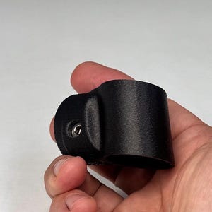 May include: A black, cylindrical, 3D-printed plastic clamp featuring a protruding side mount with a single integrated metal screw. The component has a textured surface finish and a hollow center, designed for securing cylindrical objects.