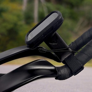 May include: Black Garmin cycling computer mounted on black bicycle handlebars. The computer is rectangular with a screen. The handlebars are wrapped in black tape. The background is blurred, showing a green and brown landscape.