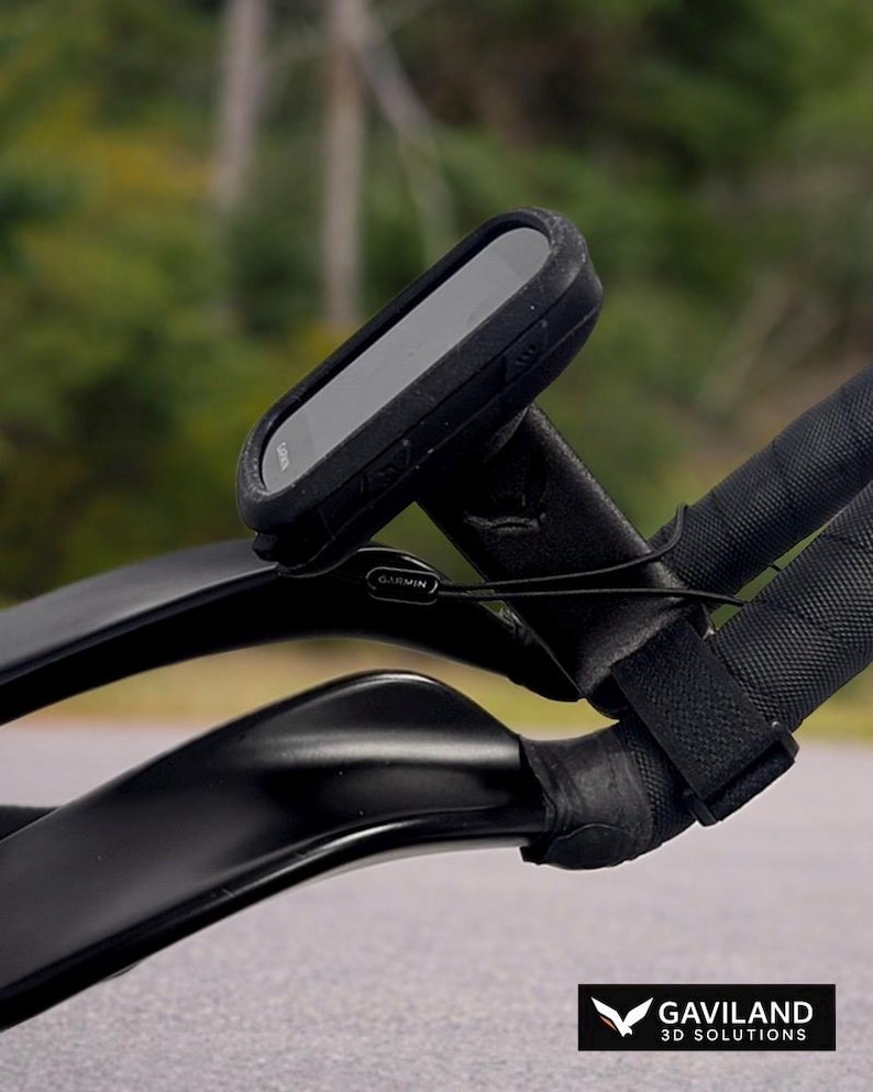 May include: Black Garmin cycling computer mounted on black bicycle handlebars. The computer is rectangular with a screen. The handlebars are wrapped in black tape. The background is blurred, showing a green and brown landscape.