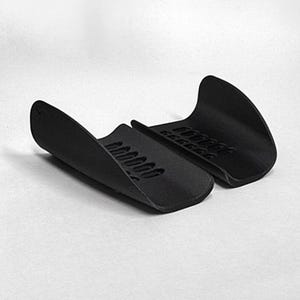 May include: A pair of matte black, curved, semi-rigid protective guards featuring a series of rectangular ventilation slots along the center. The items are positioned side-by-side, displaying a smooth, contoured exterior surface and a textured interior.