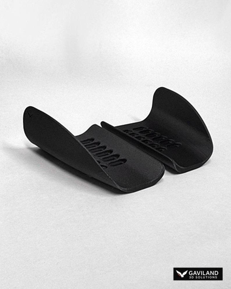 May include: A pair of matte black, curved, semi-rigid protective guards featuring a series of rectangular ventilation slots along the center. The items are positioned side-by-side, displaying a smooth, contoured exterior surface and a textured interior.