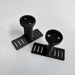 May include: Two black 3D-printed bicycle computer mounts. Each mount features a circular top with screw holes, a vertical support, and a rectangular base with three slots. The mounts are on a white surface.