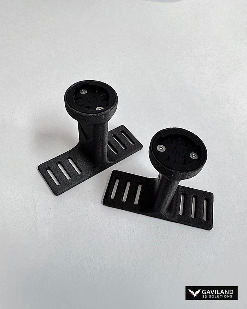 May include: Two black 3D-printed bicycle computer mounts. Each mount features a circular top with screw holes, a vertical support, and a rectangular base with three slots. The mounts are on a white surface.