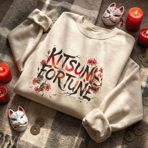 May include: Cream-colored sweatshirt with the words "Kitsune Fortune" in black and red, featuring a fox mask and floral accents. The image includes two fox masks, candles, and coins.