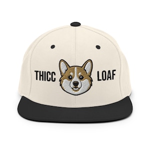 May include: A cream-colored flat-brimmed baseball cap with a black brim and top button. The cap features the words "THICC LOAF" in black block letters, with a cartoon corgi dog in the center.