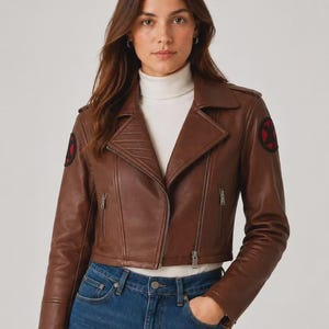 May include: A brown leather motorcycle jacket featuring a wide collar, silver-toned metal zippers, and circular black patches with a red X symbol on each shoulder. The jacket is worn over a white turtleneck sweater and blue denim jeans.